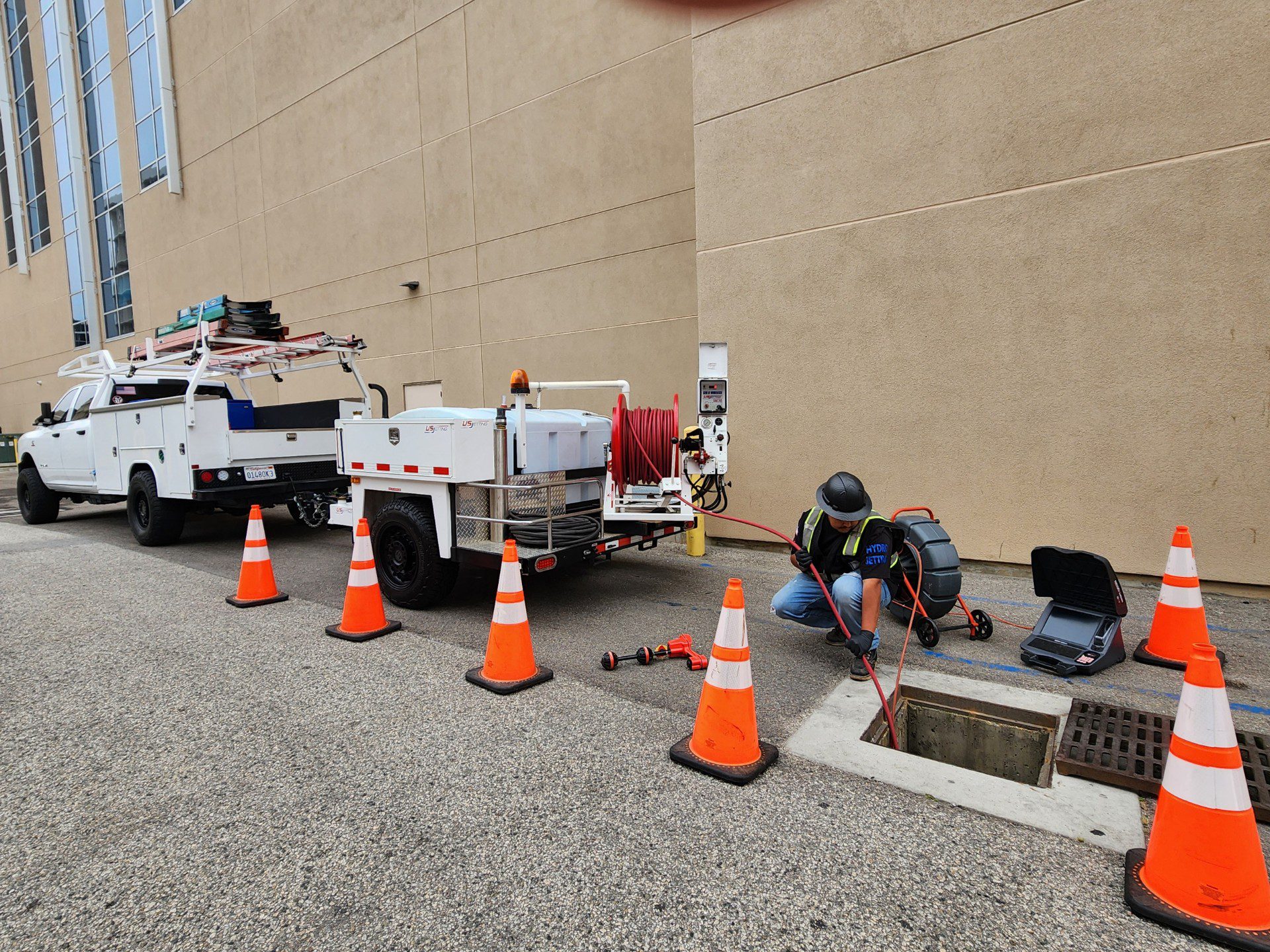 Armor service truck and jetter trailer parked behind safety cones while a technician runs a camera line into a commercial storm drain