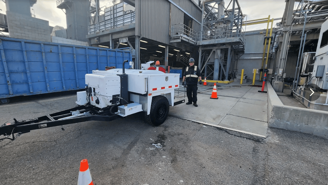 Hydro jetting trailer from Armor Plumbing & Hydro Jetting set up on an industrial site with a crew member in a safety vest standing beside it